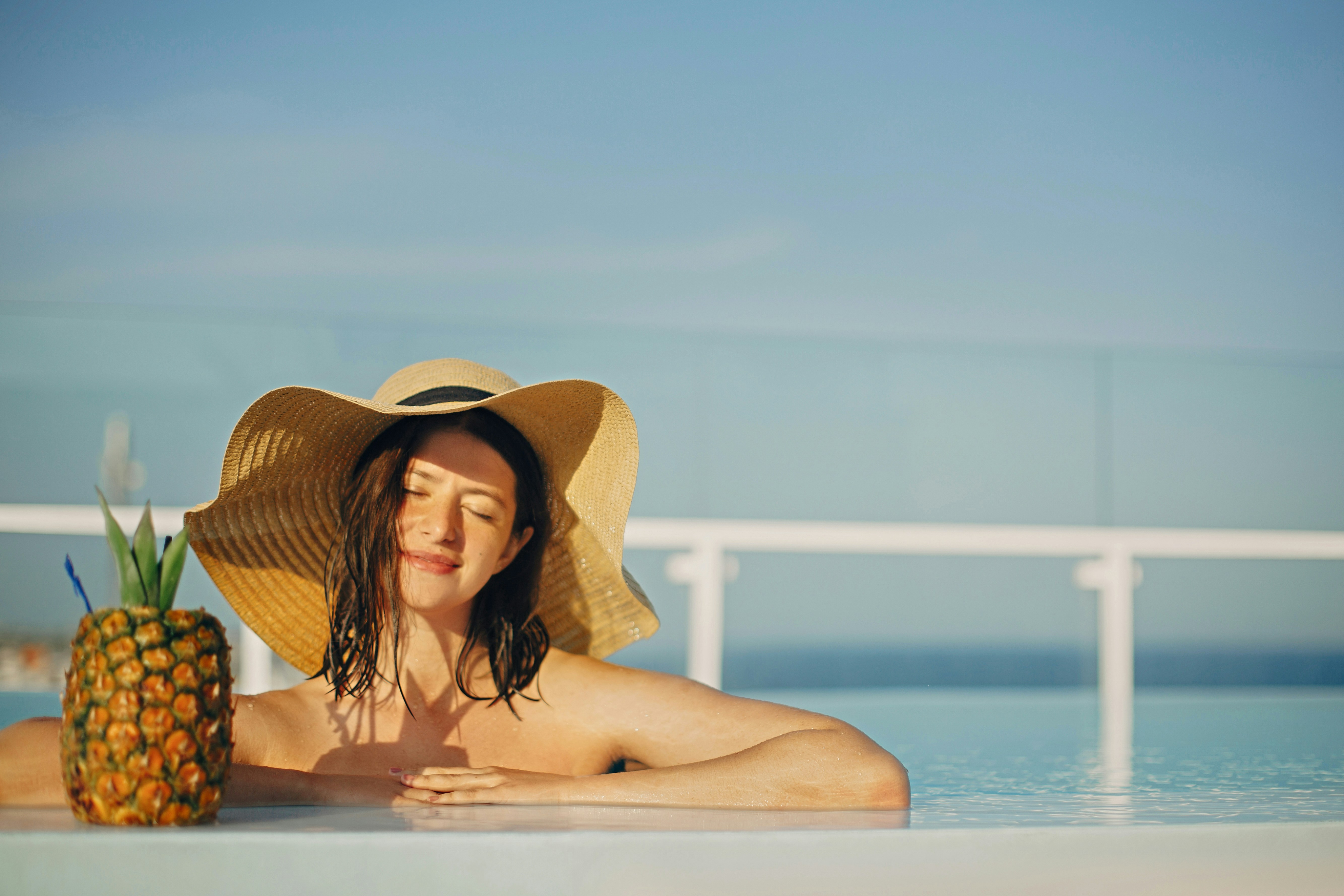 Woman relaxing in a pool with a pineapple drink, wearing a sun hat