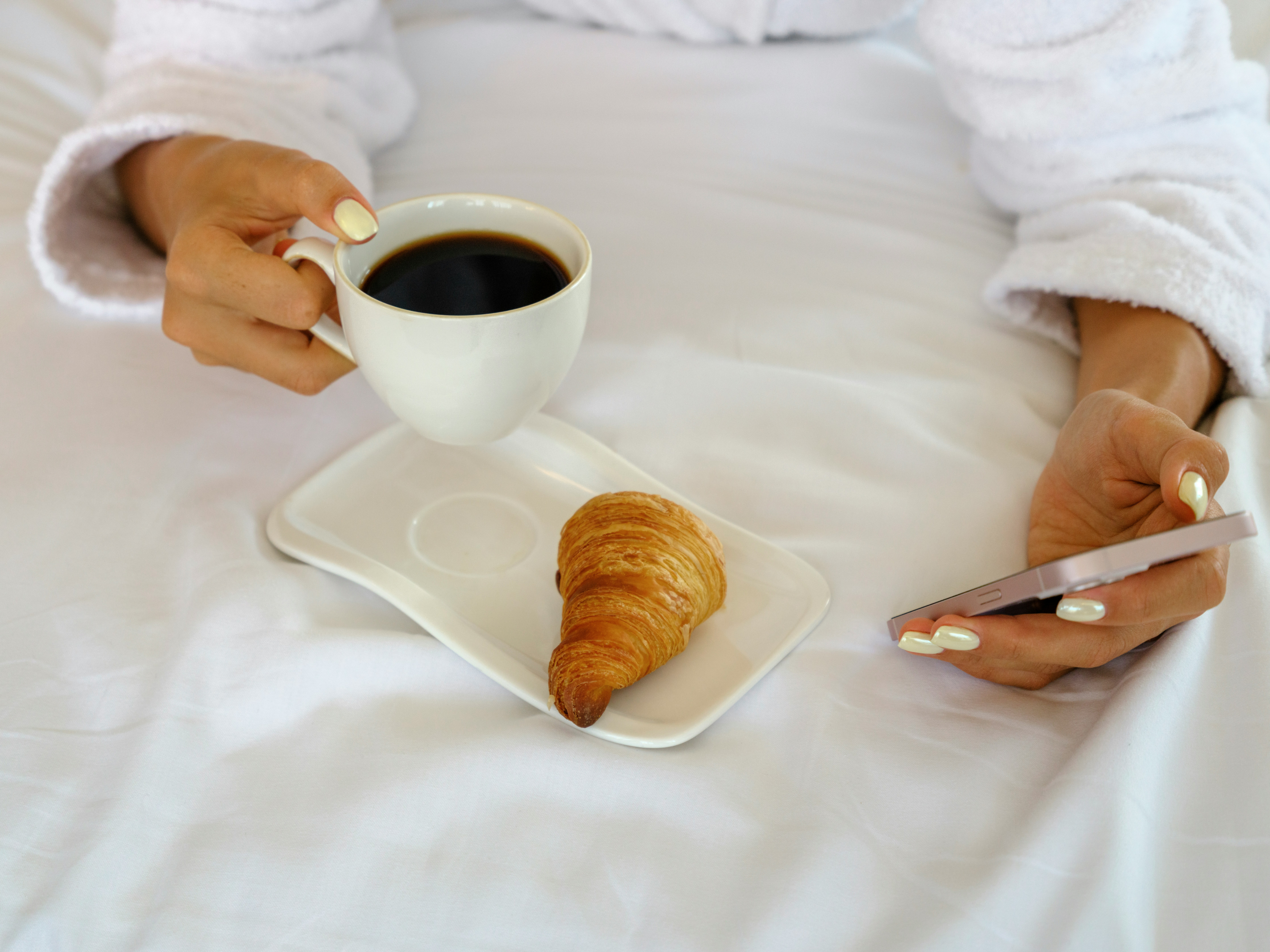 Person relaxing in bed with coffee and a croissant, using a smartphone